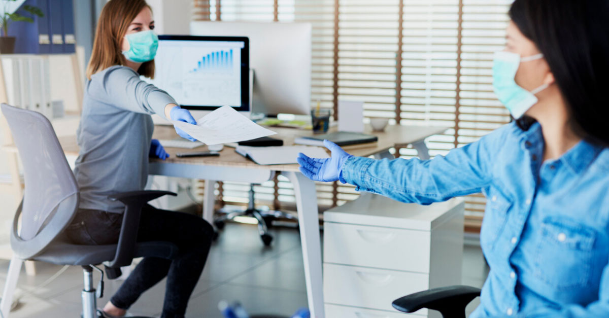 Two employees at a doctor's office working in an office wearing masks, handing paper to one another.